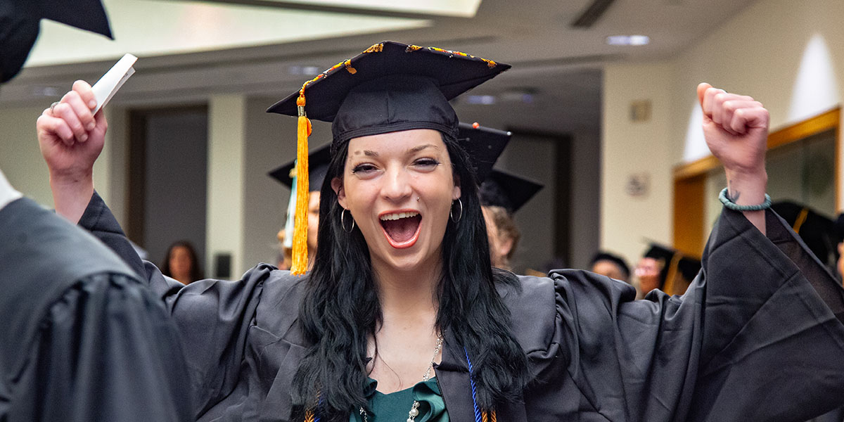 An NMC graduate in cap and gown raising her hands and smiling during NMC's 2020 Commencement